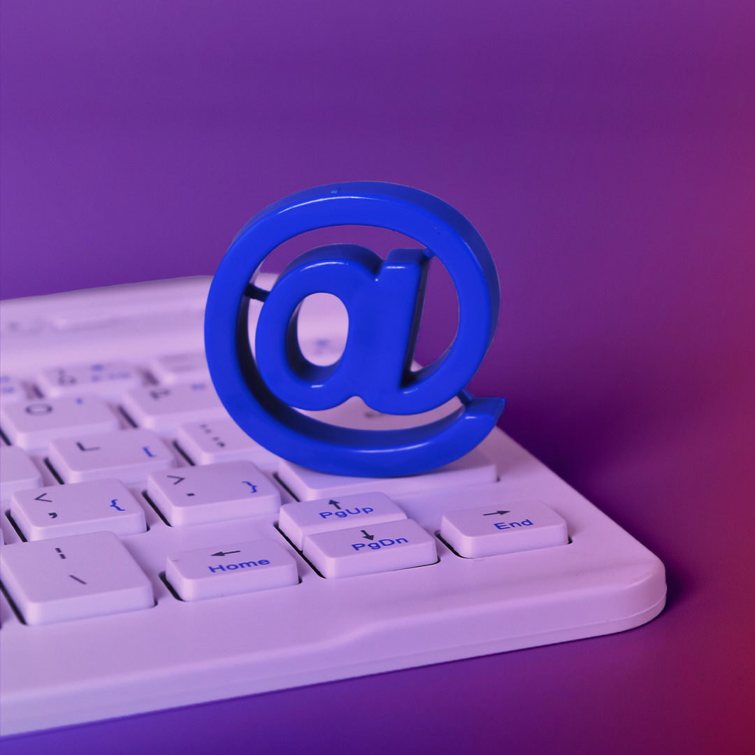 A blue at-symbol figurine is placed on the corner of a white computer keyboard resting on a purple surface. OCIDM,io Branding and Digital marketing Hamilton, Toronto, Oakville, Mississauga