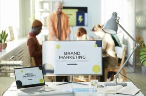 A desk with a monitor and laptop displaying "Brand Marketing." A desk lamp is on the right. In the background, four people are having a meeting focused on brand positioning in a well-lit room. OCIDM,io Branding and Digital marketing Hamilton, Toronto, Oakville, Mississauga