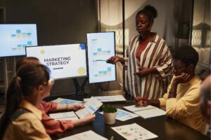 In a dimly lit meeting room, a woman presents an SEO-focused marketing strategy to three colleagues, with screens displaying graphs. OCIDM,io Branding and Digital marketing Hamilton, Toronto, Oakville, Mississauga