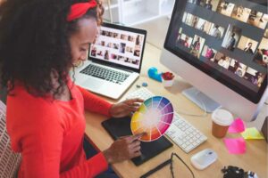 A person sits at a desk using a color wheel with a laptop and monitor displaying images. Nearby, tools for a brand-intensive project—like a camera, coffee cup, and sticky notes—are scattered around as they focus on enhancing SEO strategies. OCIDM,io Branding and Digital marketing Hamilton, Toronto, Oakville, Mississauga