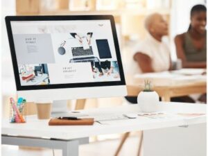 A desktop computer showcasing professional web design software sits on a desk adorned with office supplies, a notebook, and a potted plant. In the blurred background, two business professionals engage in conversation at a table in Toronto. OCIDM,io Branding and Digital marketing Hamilton, Toronto, Oakville, Mississauga